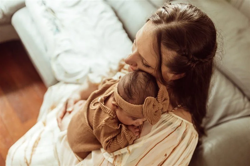 A woman holding a sleeping baby on her shoulder on a beige couch.
