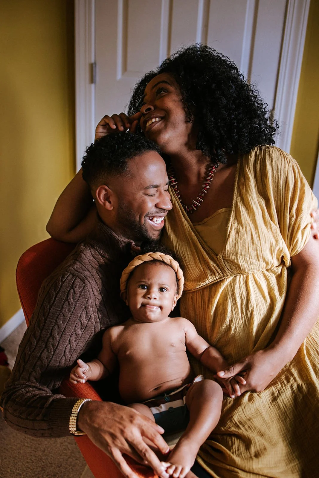 A smiling family of three with a baby sitting on the father's lap, the mother is standing and touching her face, all sharing a joyful moment in a cozy room.