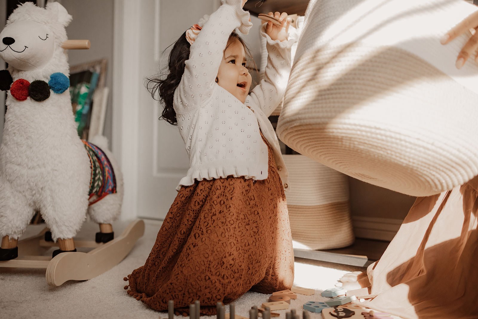 A young girl with dark hair, wearing a cream-colored cardigan and a rust-colored skirt, kneels on a carpeted floor, playing with toys. There is a plush llama on wheels on her left and stacked baskets behind her to the right.