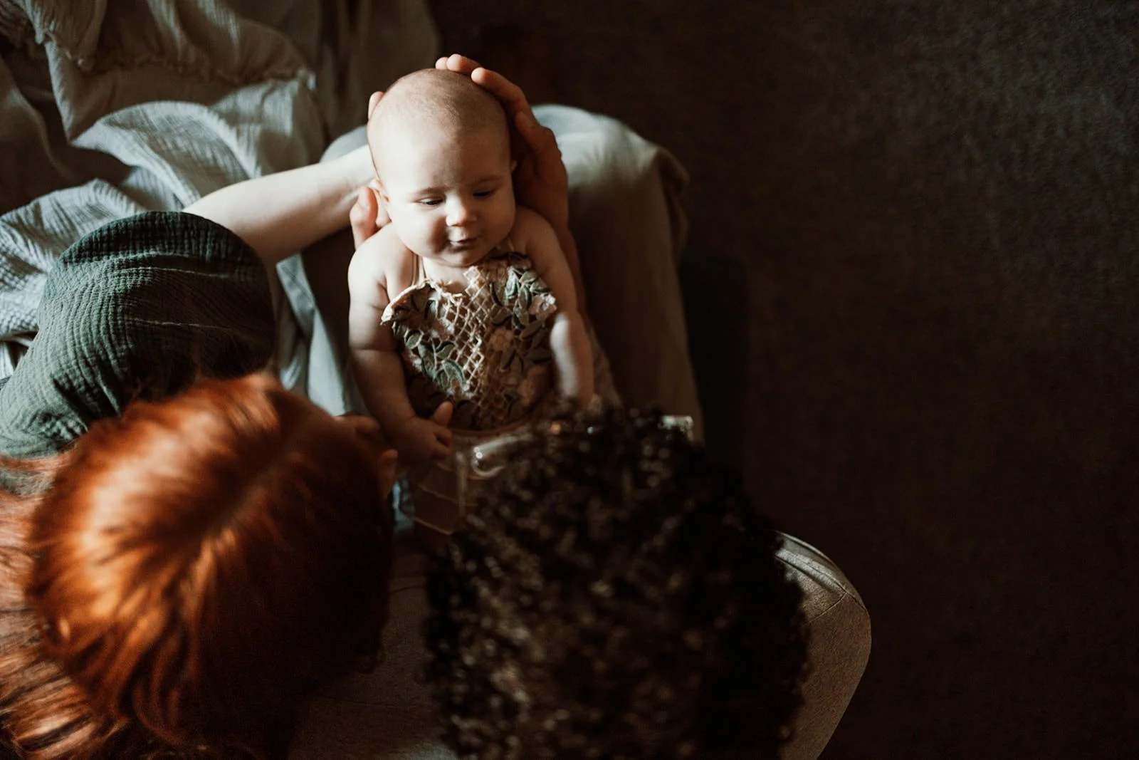 A baby with a patterned dress sitting on a bed, being held by an adult, while another person with reddish hair hands a small gift box to the baby. The scene is dimly lit, creating a cozy atmosphere.