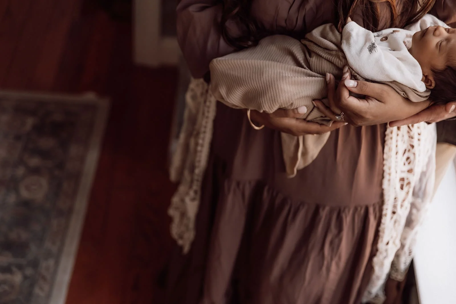 A woman holding a sleeping baby wrapped in a blanket in her arms, with only her hands visible, wearing a ring and a bracelet, standing near a table with a patterned rug underneath.
