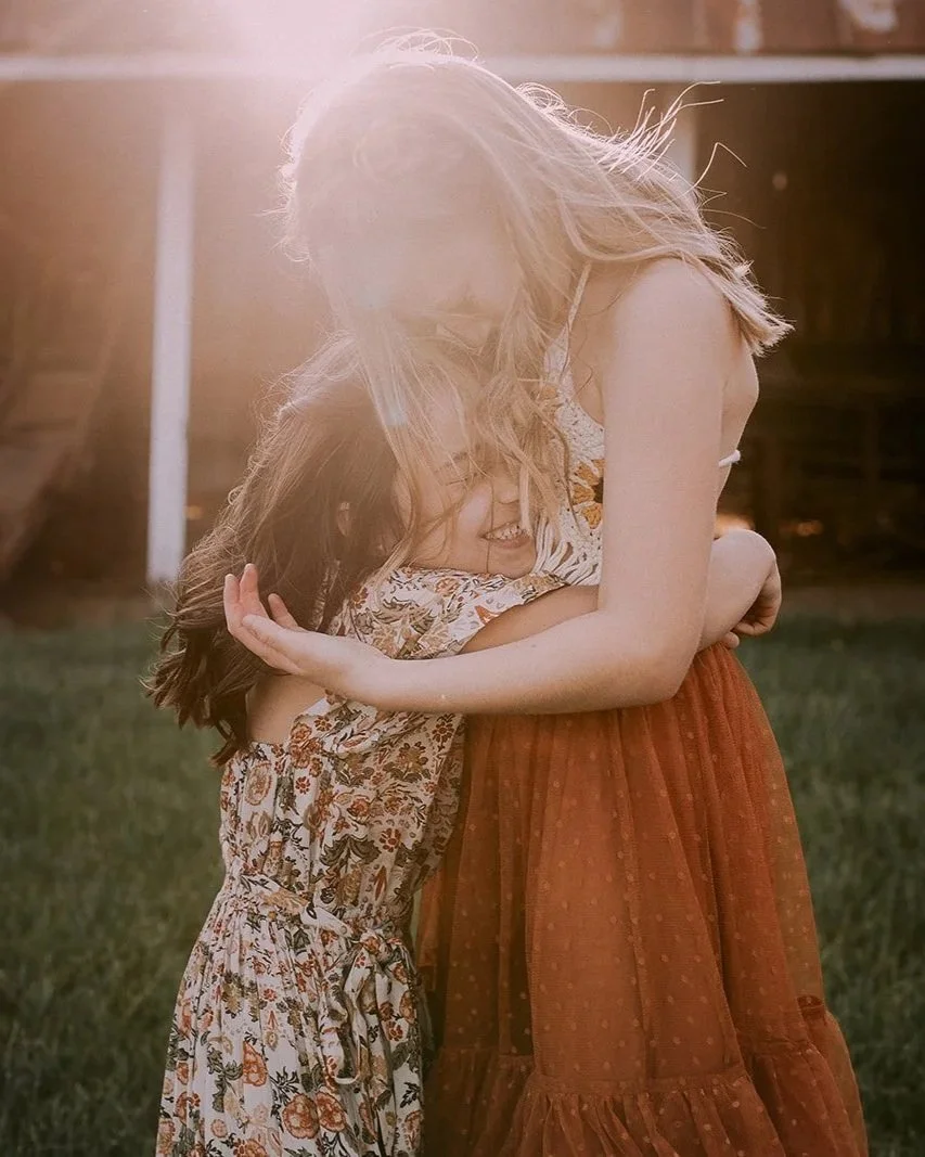 Two girls hugging outdoors with sunlight in the background, one with long wavy hair wearing a patterned dress, and the other with long straight hair wearing a reddish-orange skirt.