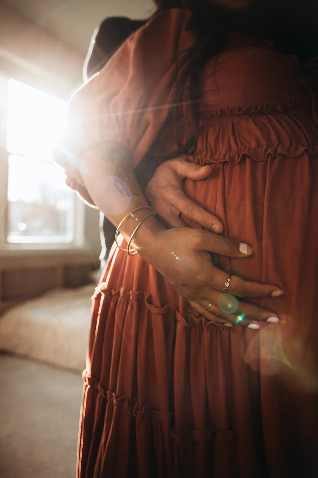 A pregnant woman wearing a rust-colored dress is holding her belly, with sunlight coming through a window behind her.