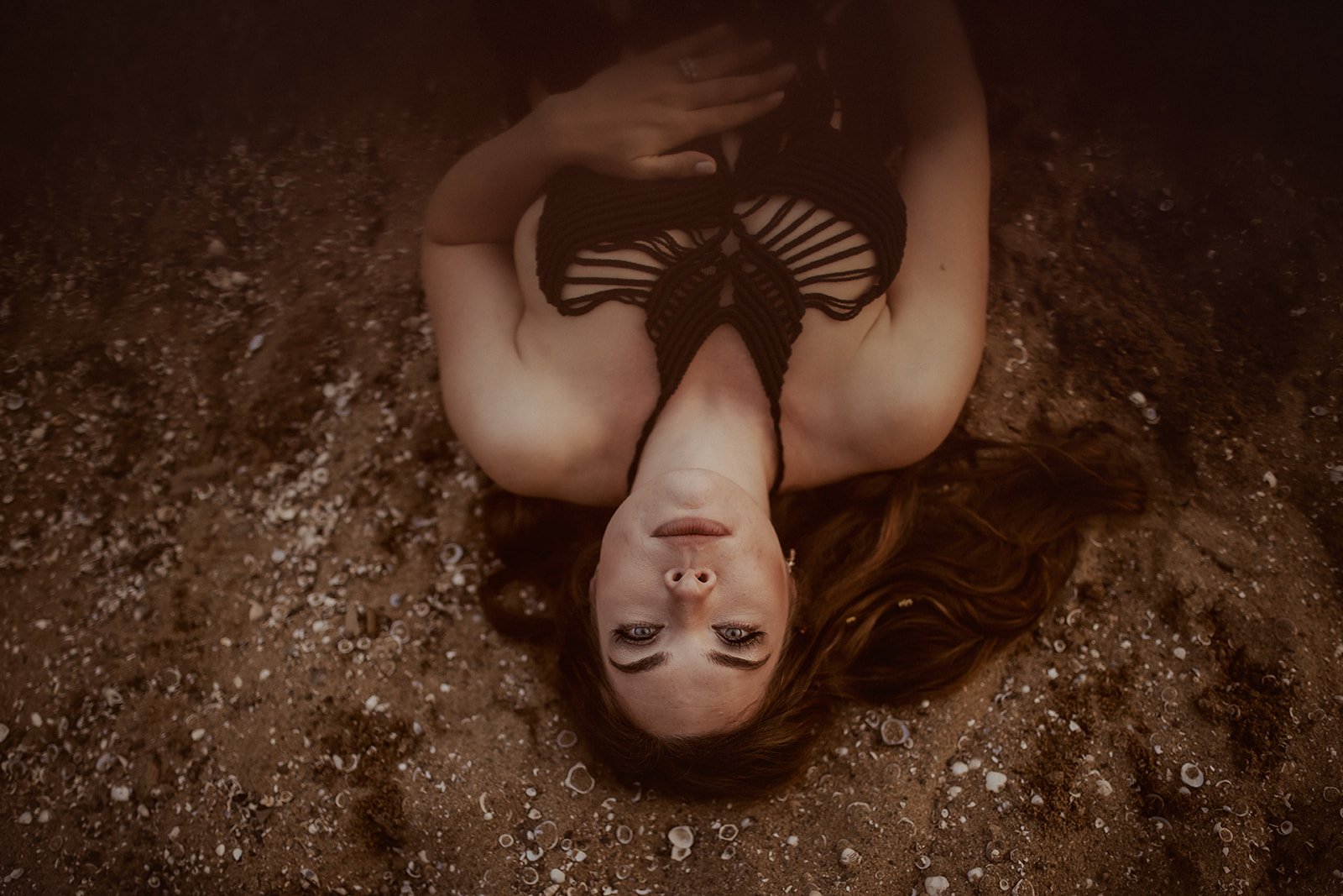 A woman with long brown hair lying on a sandy surface, wearing a black, intricately designed halter top, with her eyes open and looking at the camera.