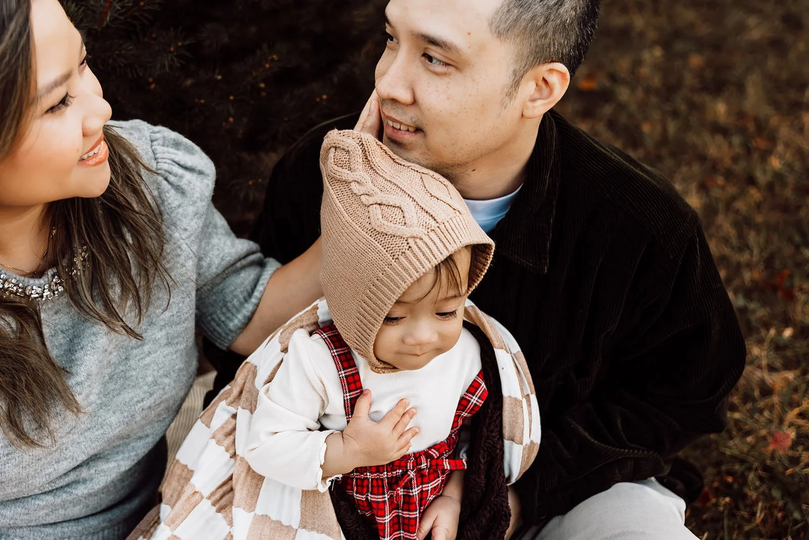 A family of three sitting outdoors during autumn; a woman and man looking lovingly at each other and their young child, who is wearing a beige knit hat with a pom-pom and a plaid jumper, while she holds her hand over her chest.