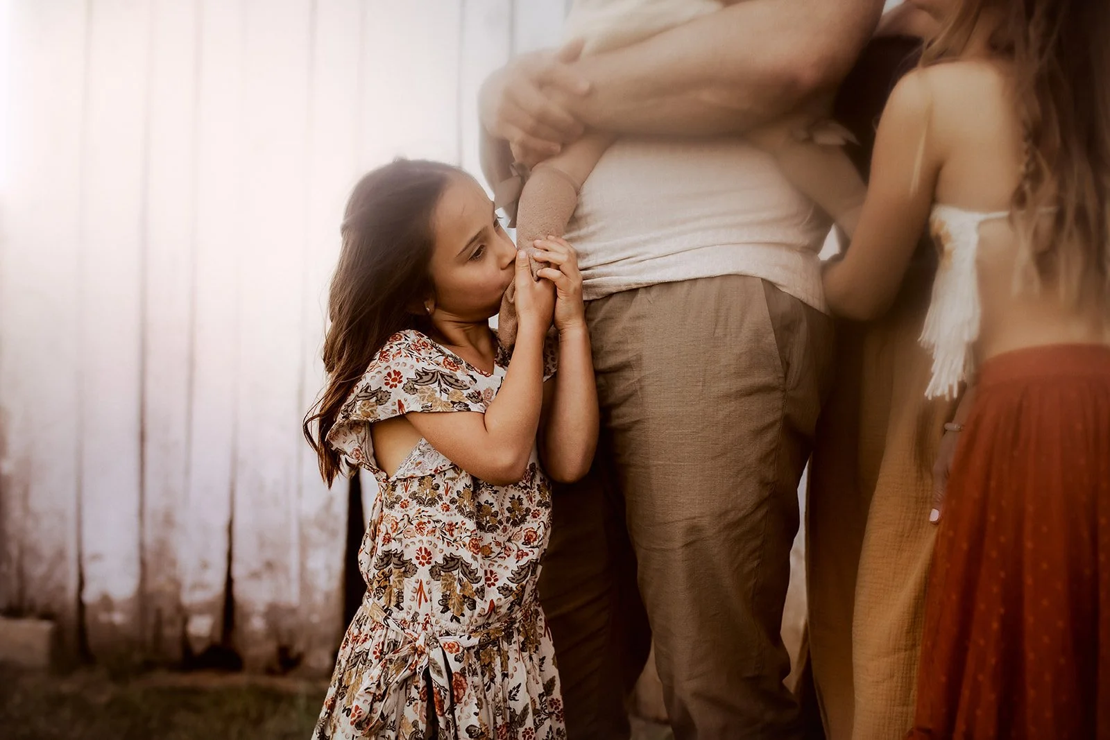 A young girl with long brown hair wearing a floral dress, kissing a man's arm, while two women in bohemian-style clothing stand nearby.