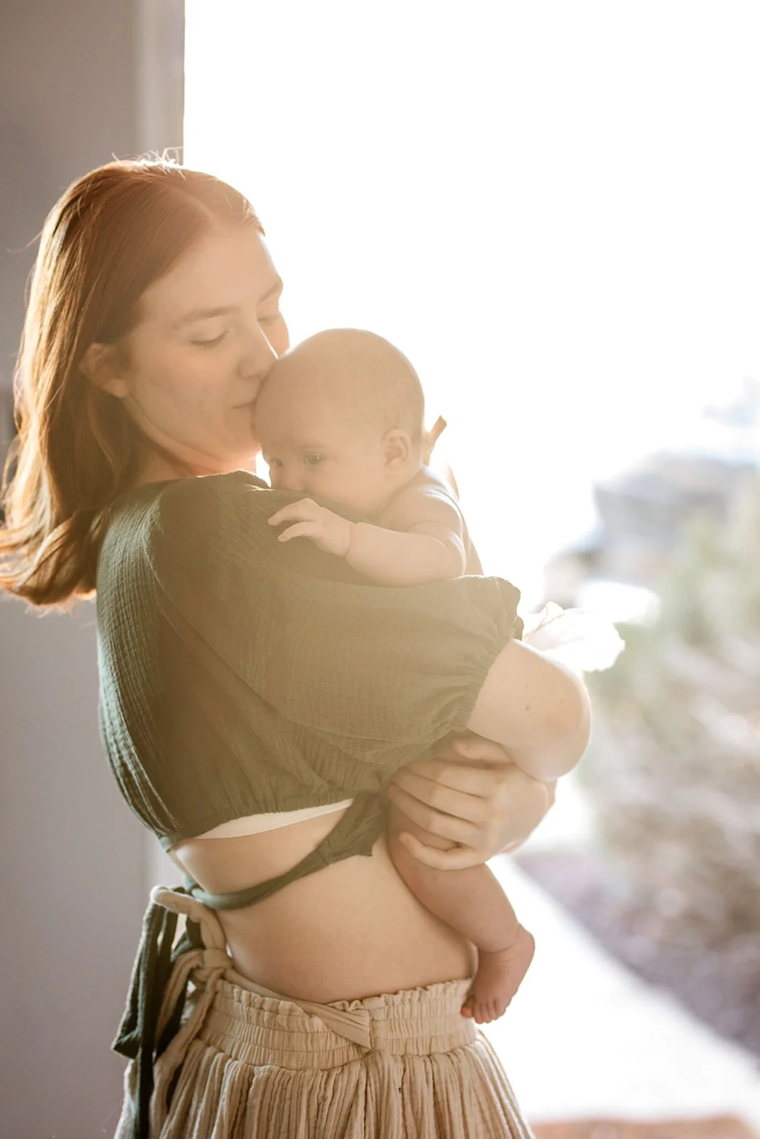 A red-haired woman holding a baby, gently kissing the baby's forehead indoors near a bright window.