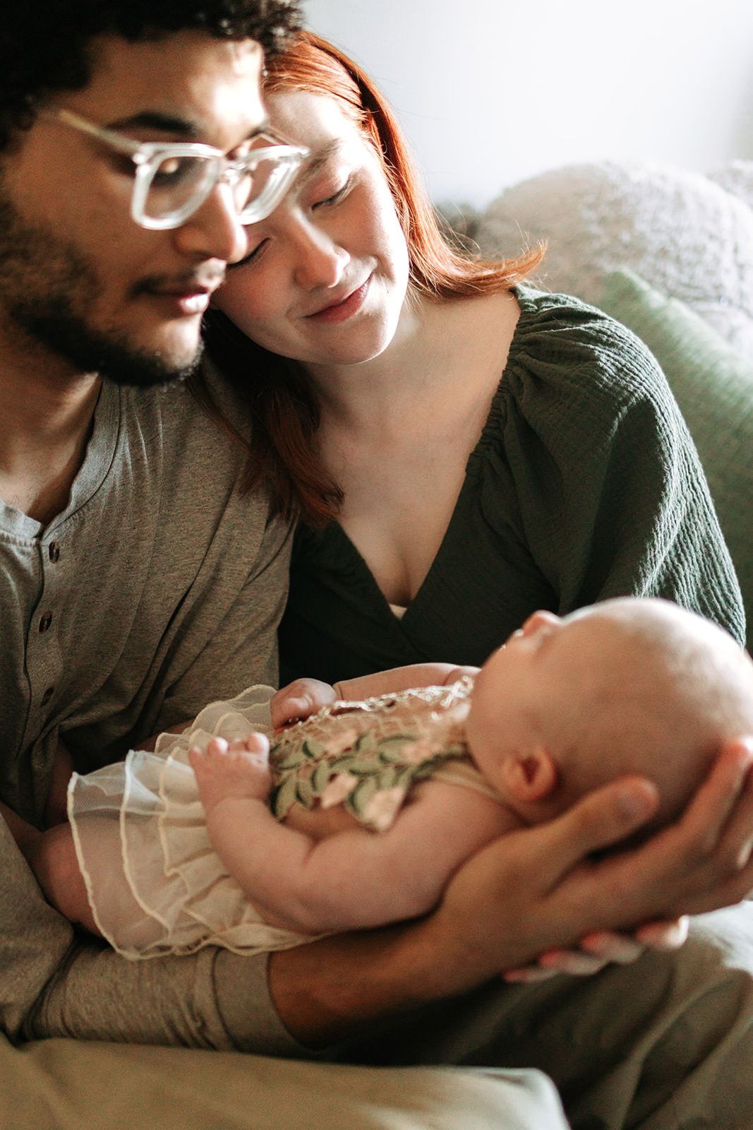 A young couple holds a baby, with the woman smiling and looking down at the infant, while the man looks at the baby with glasses on. The baby has a floral outfit and is looking up.