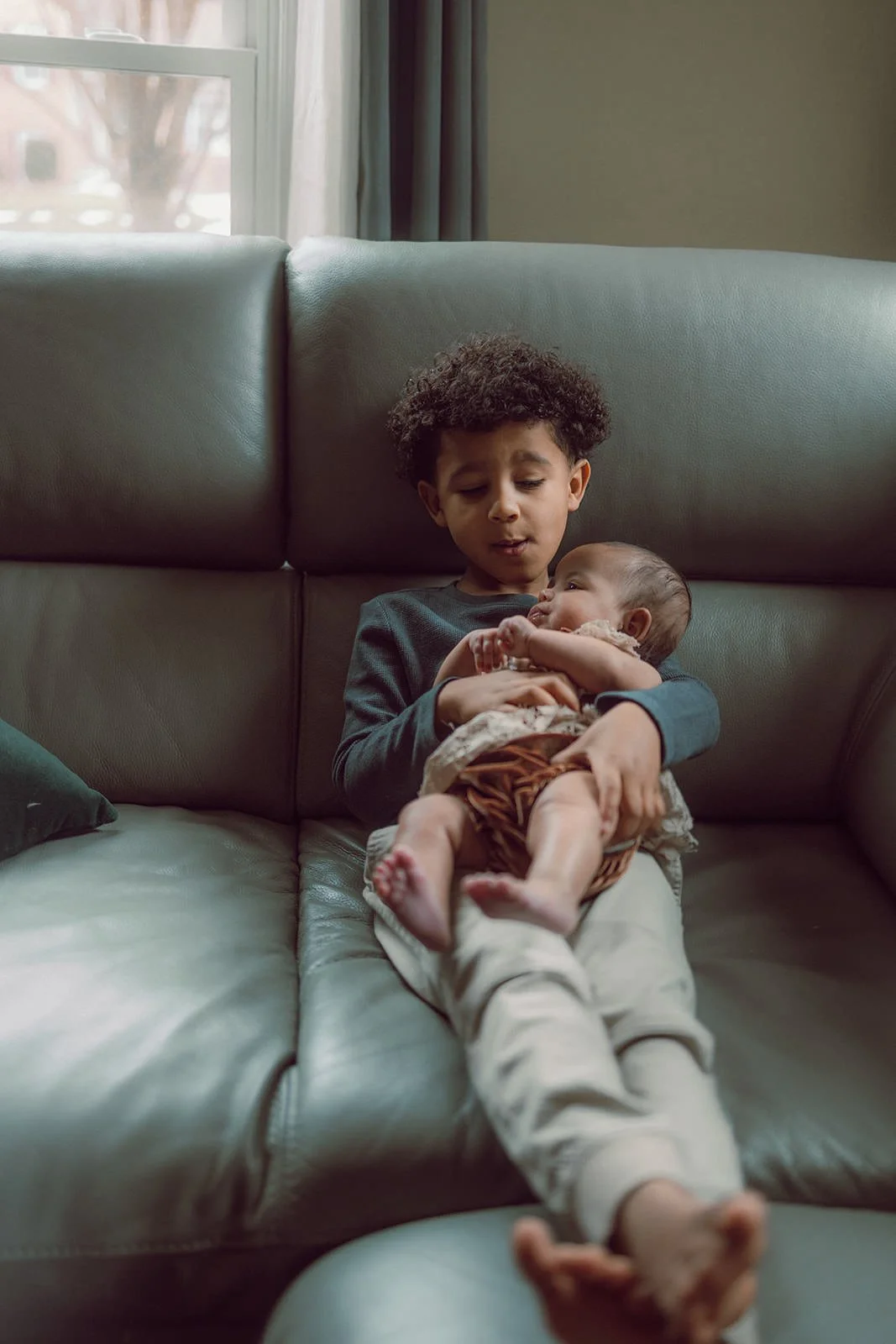 A young boy sitting on a sofa holding a newborn baby in his arms.