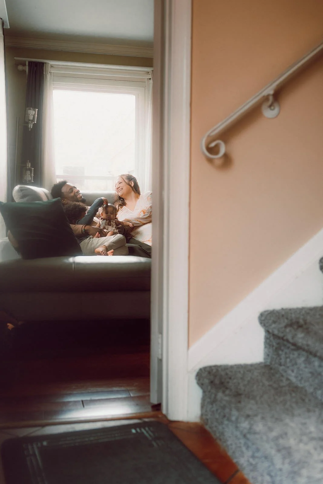 A family sitting on a couch in a living room, smiling and enjoying each other's company, viewed through a doorway or staircase.