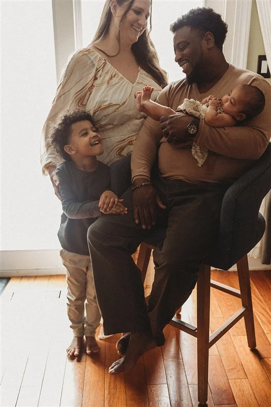 An African American family of four, including a man holding a newborn baby girl, a smiling woman, and a young child, gathered indoors on a wooden floor, sharing a joyful moment.