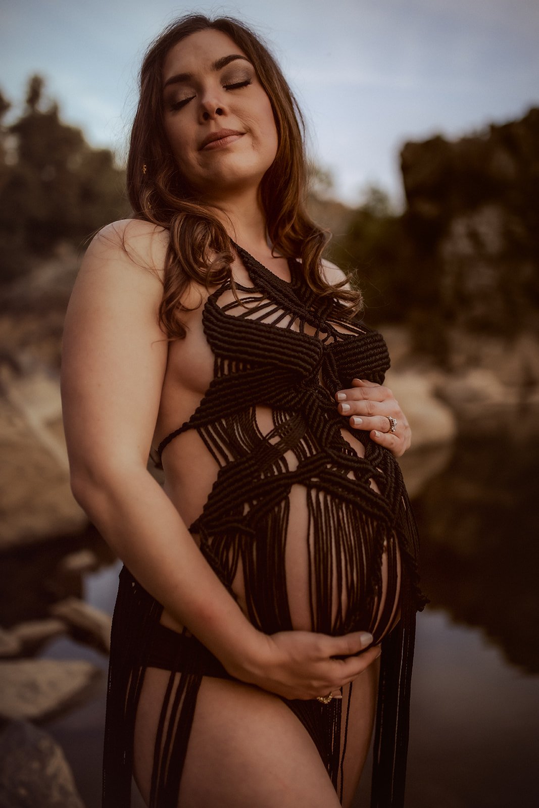A pregnant woman with light skin and long brown hair, wearing a black, open, stringy dress, standing outdoors near rocks and water at sunset, with her eyes closed and one hand on her belly.