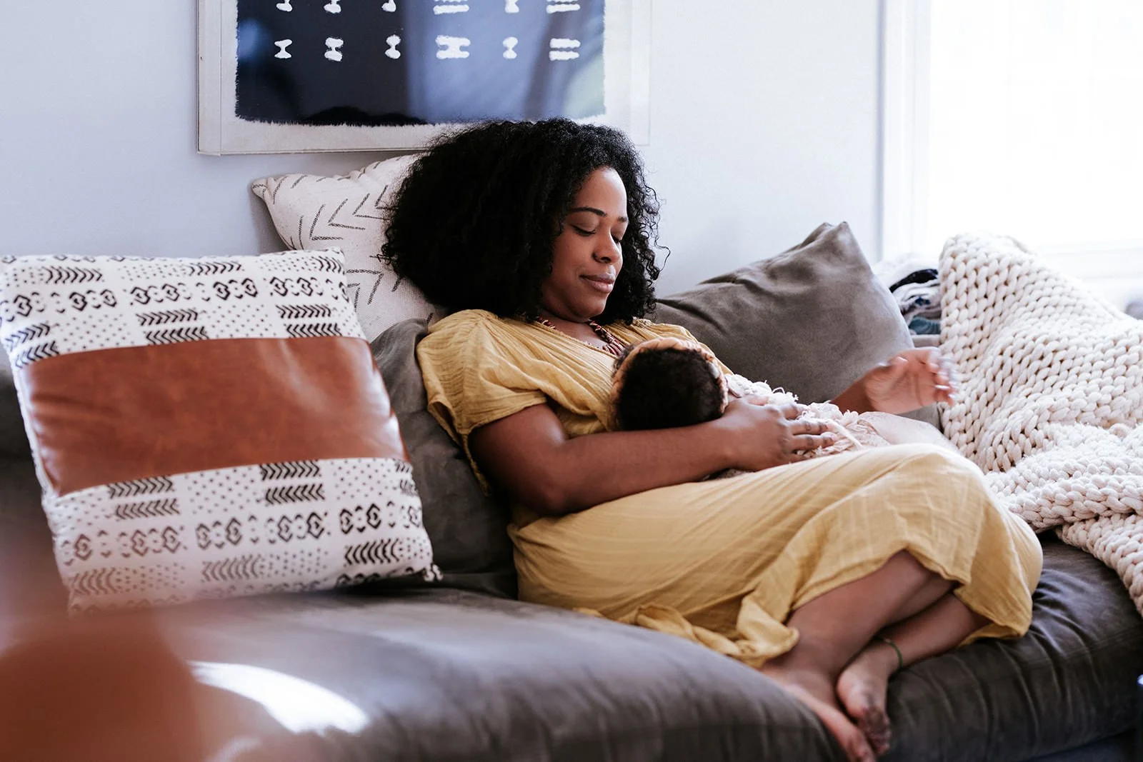 A woman with dark curly hair in a yellow dress is sitting on a couch, breastfeeding a baby. The couch has several decorative pillows and a chunky knit throw blanket. There is abstract wall art in the background and natural light coming from a window.