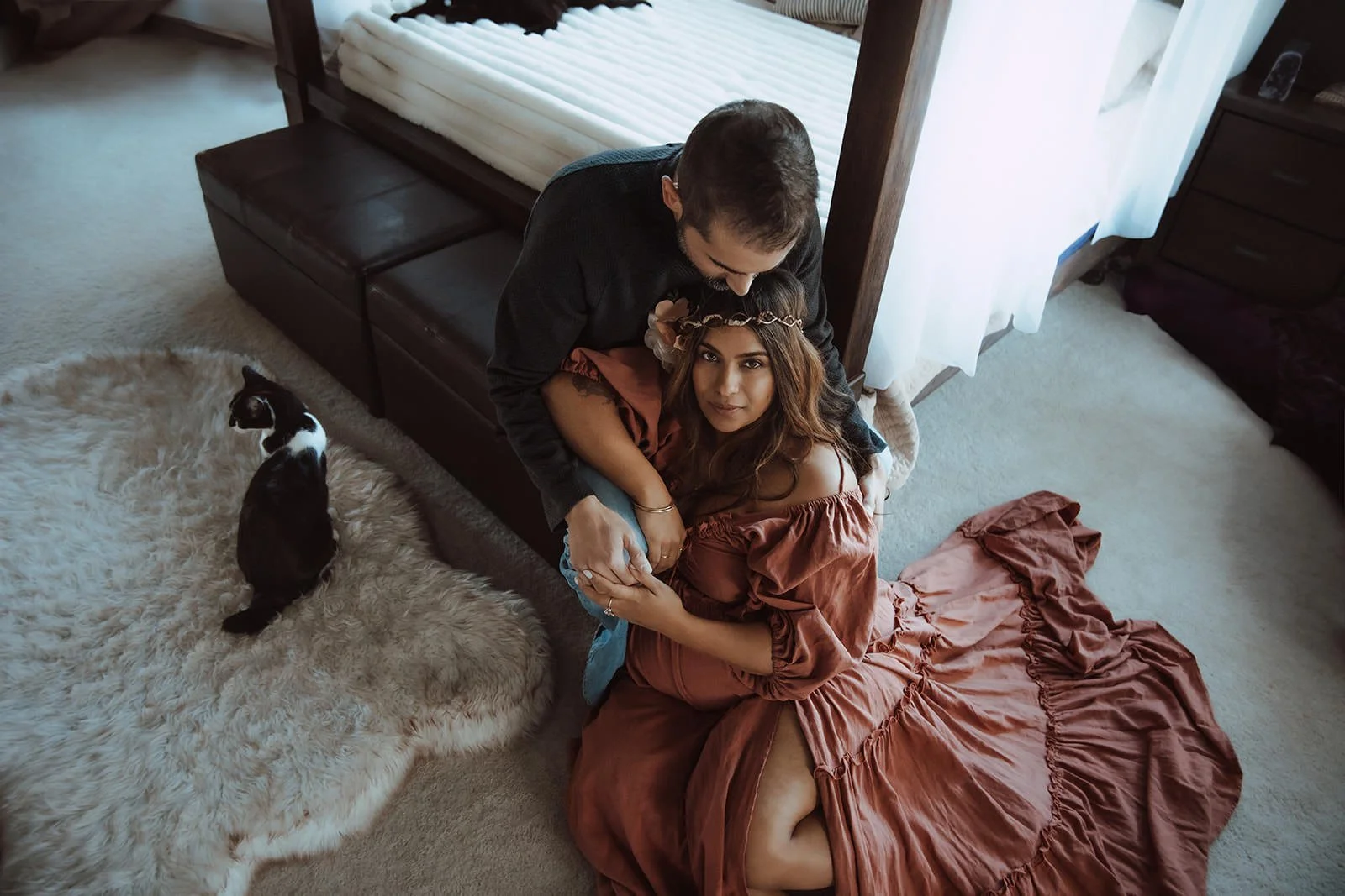 A couple is sitting on the floor inside a room, with the woman wearing a long, ruffled dress and a floral crown, looking up at the camera while the man leans over her. A black and white cat sits on a fluffy rug nearby. The room has natural lighting c