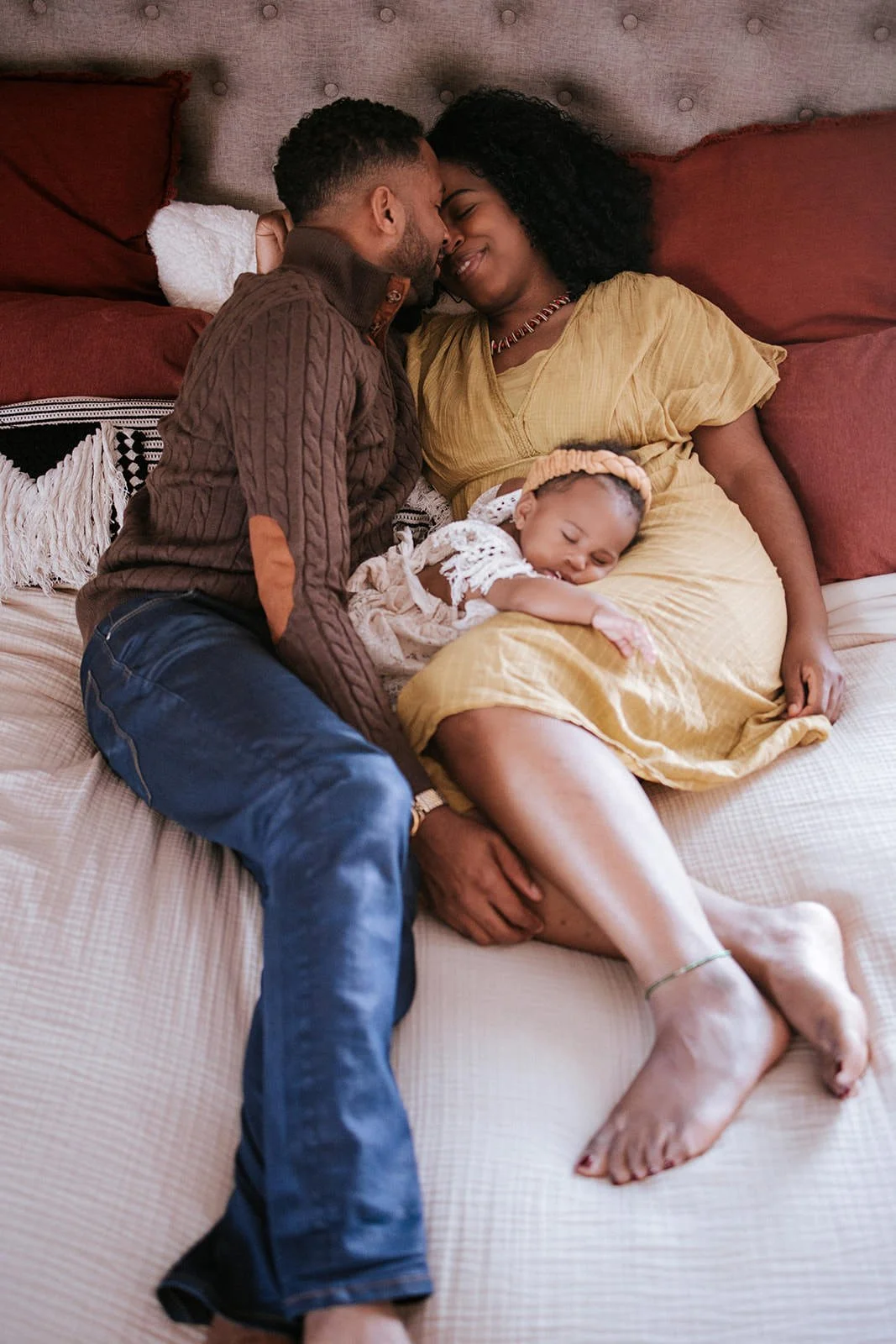 A family of three lying on a bed, with a pregnant woman in a yellow dress smiling, a man leaning close with eyes closed, and a small girl resting on the woman's belly with her eyes closed.