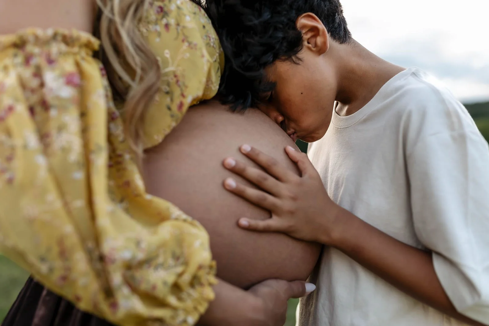 A pregnant woman with a yellow floral dress and a young man wearing a white shirt sharing an intimate moment, with the man kissing her pregnant belly.