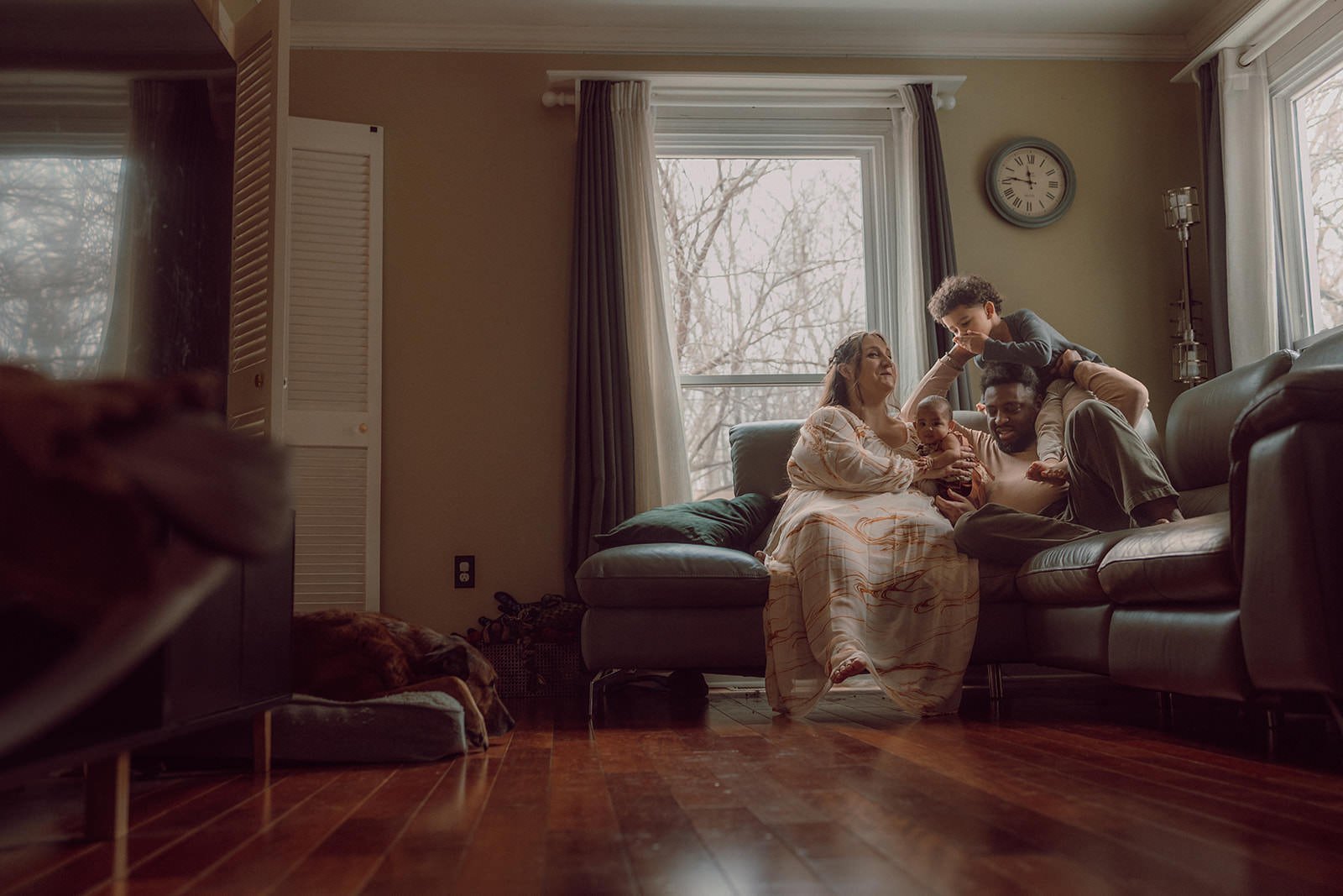 A family of four sitting on a sofa in a living room, with a child playing on the man's shoulders. The mother and baby are sitting on the sofa, and a dog is sleeping on a bed near the camera.