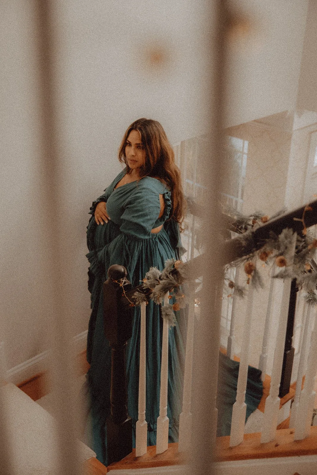 A woman in a teal dress with puffed sleeves standing on a staircase decorated with flowers and greenery, looking over her shoulder.