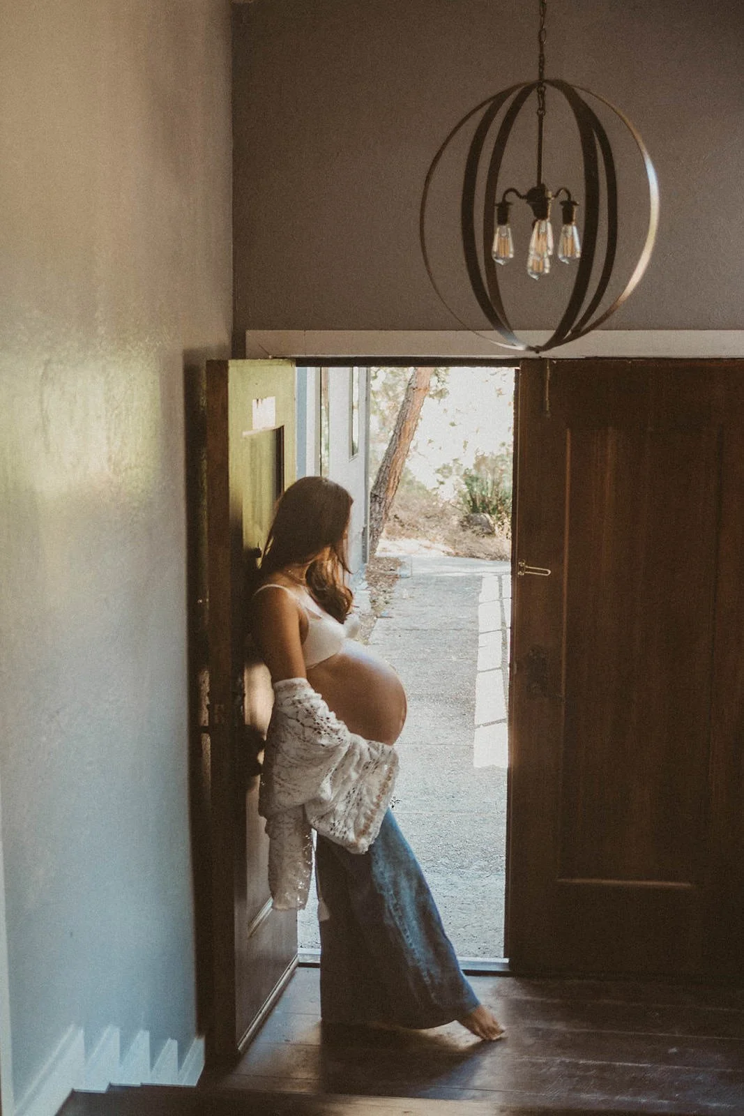 Pregnant woman standing in an open doorway, looking outside. She is dressed in casual jeans and a white top, with a white lace cardigan. The setting appears to be inside a home with a modern pendant light hanging from the ceiling.