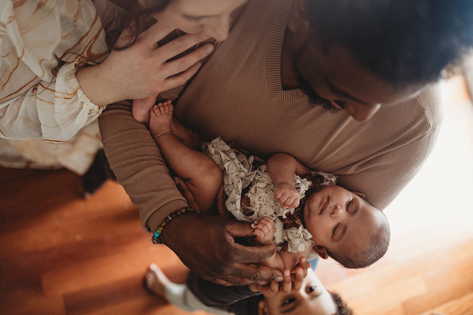 A man holding a sleeping baby girl with another child peeking from behind him, indoors with wooden flooring.