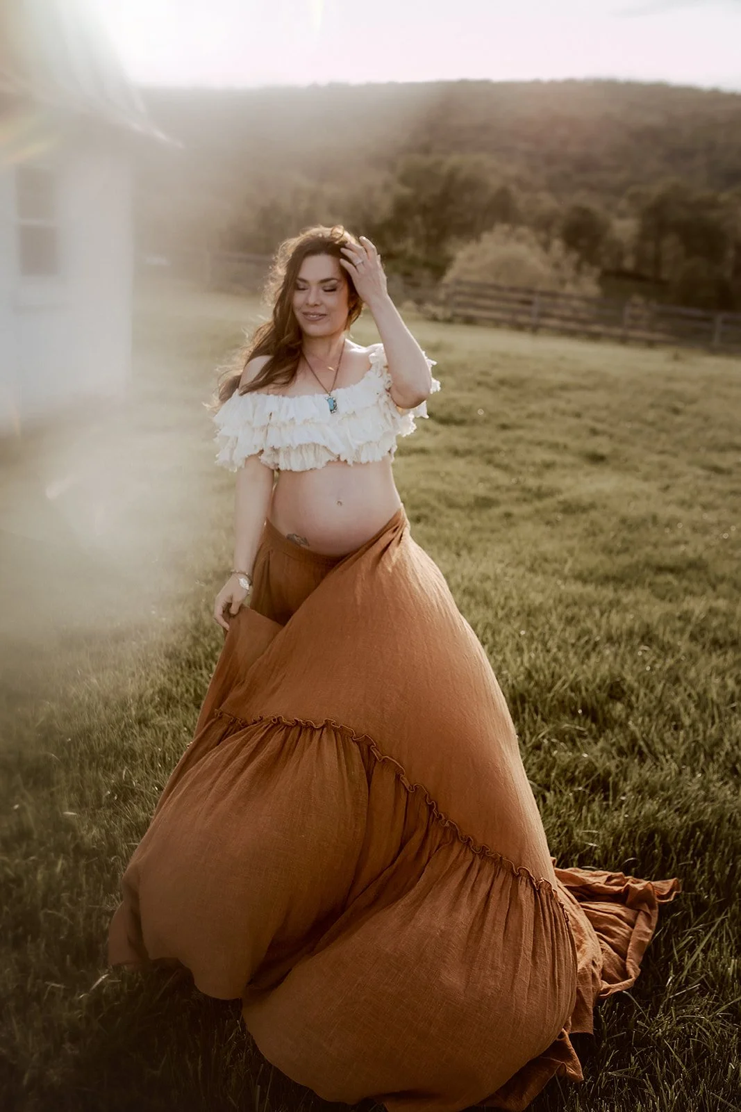 A pregnant woman with long brown hair standing in a grassy field during sunset, wearing a white off-shoulder top and a long, brown skirt.