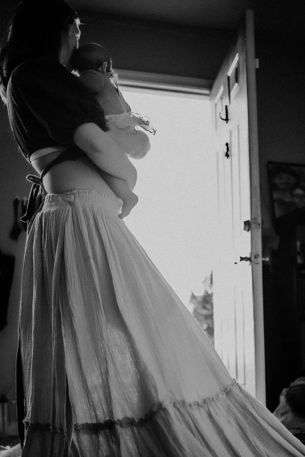A woman holding a small child near an open door, standing inside a room with sunlight streaming in, captured in black and white.