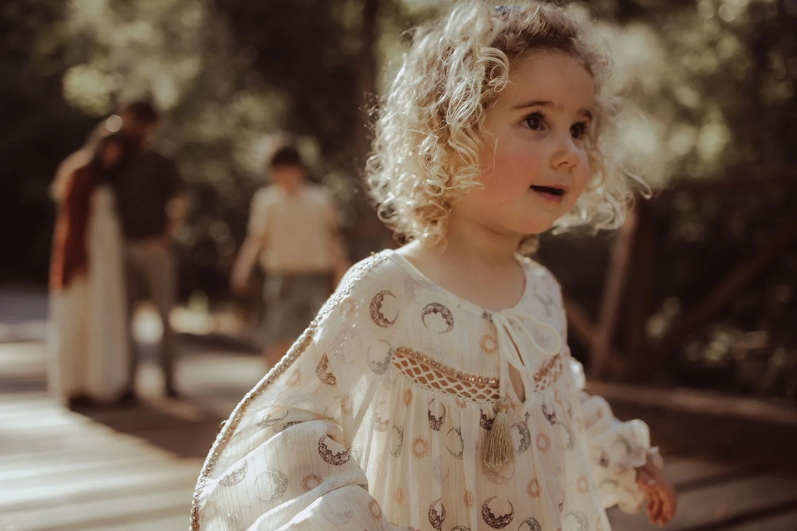 A young girl with curly blonde hair wearing a beige dress with moon and star patterns, standing outdoors with sunlight filtering through trees, and two people in the background.
