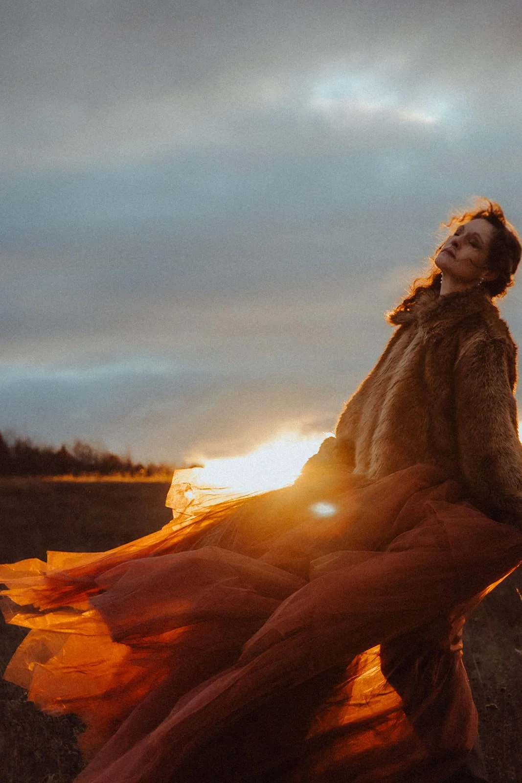 A woman with curly hair sitting outdoors during sunset, wearing a fur coat and a flowing orange skirt, with the sky and horizon in the background.
