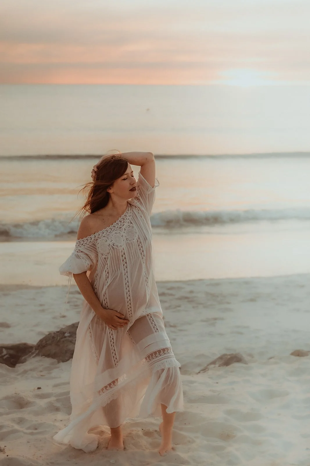 A woman with red hair standing barefoot on the sand at the beach during sunset, wearing a flowy, lace-trimmed white dress with one hand behind her head and the other on her hip.