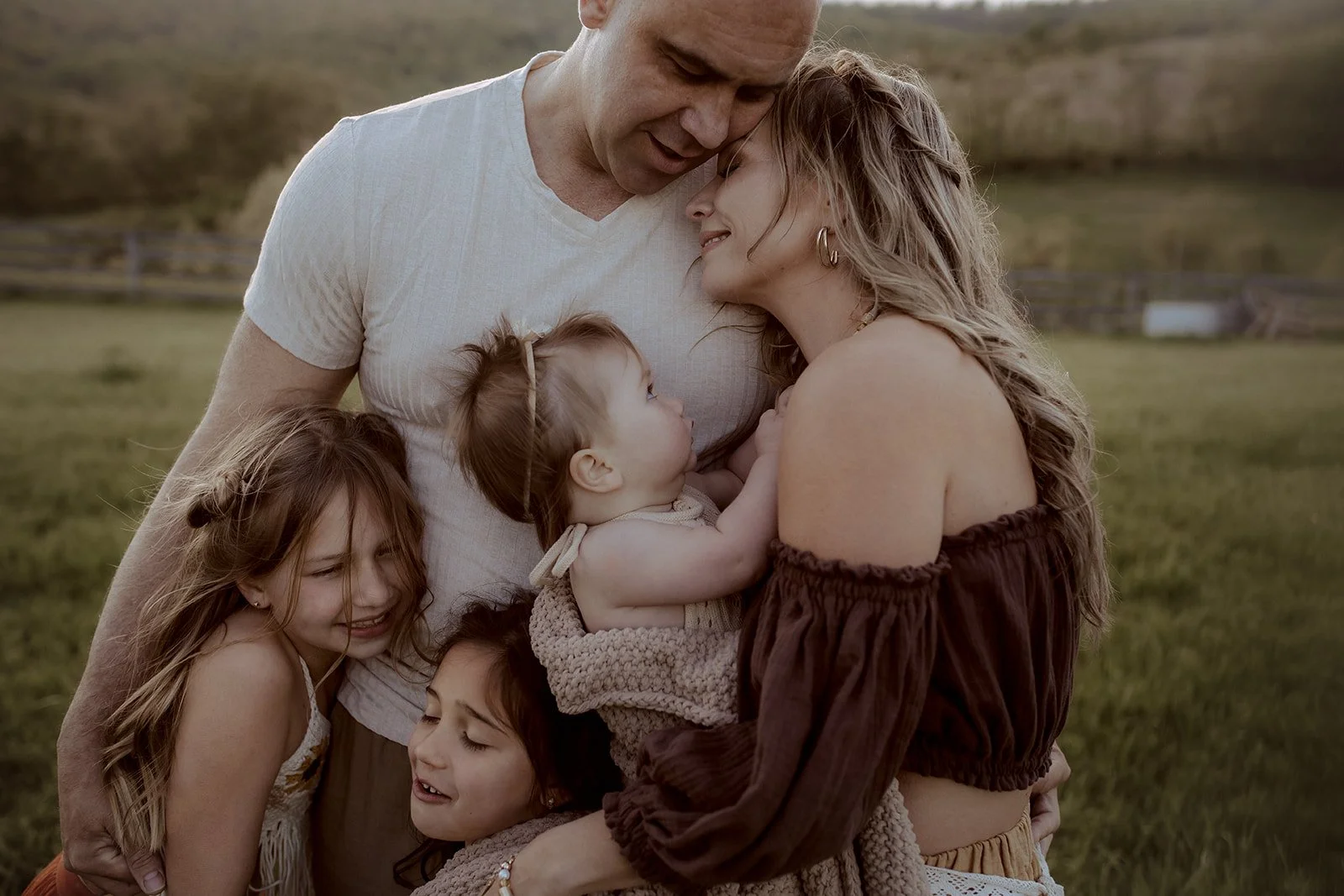 A family of six, including an adult man and woman with four young girls, is embracing outdoors on a grassy field with a fence and hills in the background. They are smiling and sharing a close, affectionate moment.