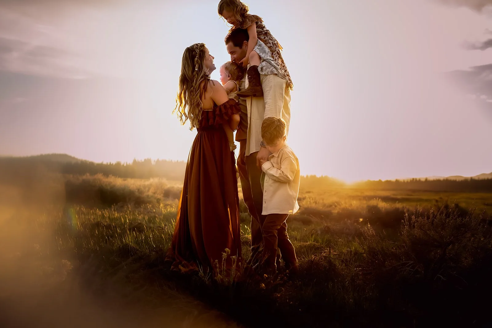 A family of six stands together outdoors during sunset, with the sun setting in the background. The mother, father, and four children are closely gathered, with the children on the father's shoulders and standing in front of the parents. The scene ap