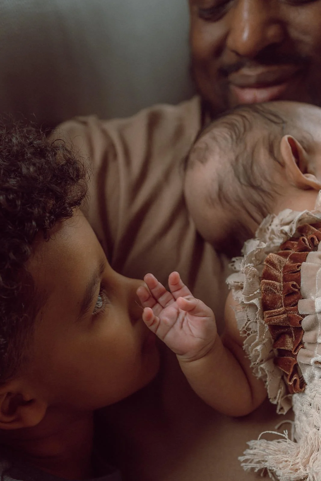 A man is holding a sleeping baby with a blanket dressed in vintage-style clothing, while a young child looks lovingly at the sleeping baby.