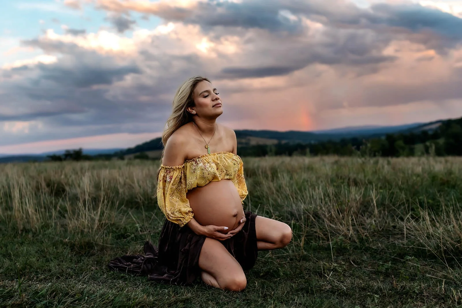Pregnant woman kneeling in a grassy field at sunset, wearing an off-shoulder yellow blouse and a dark skirt, with trees and hills in the background.