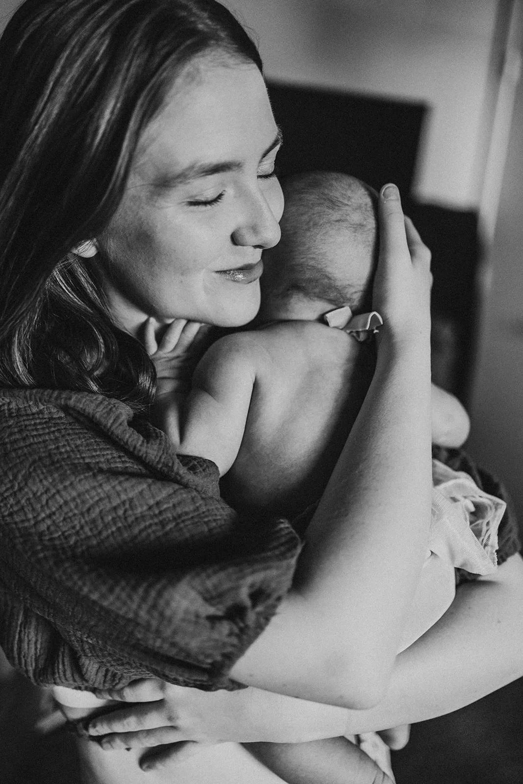 A woman with long hair, smiling with her eyes closed, embraces a small child, holding the child's head close against her cheek.