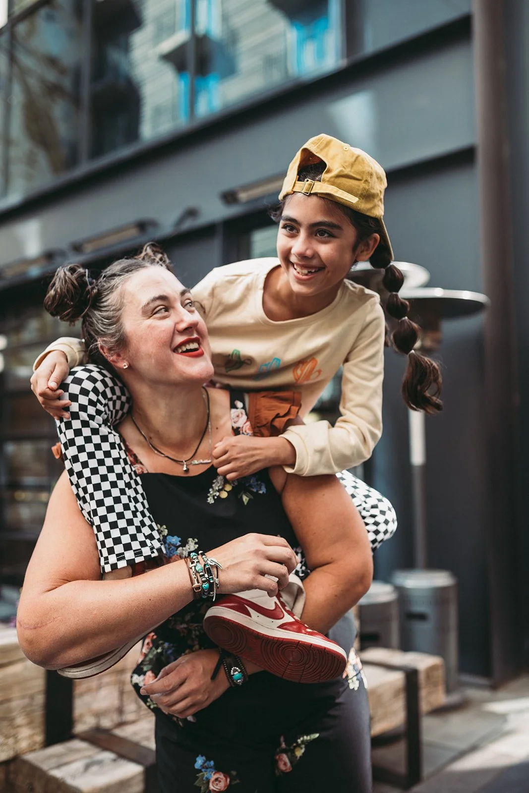 A woman with braided gray hair and colorful jewelry is carrying a smiling young girl on her shoulders inside a modern building with glass and metal decor. The girl is wearing a beige sweatshirt with colorful graphics, a yellow cap, and red sneakers.
