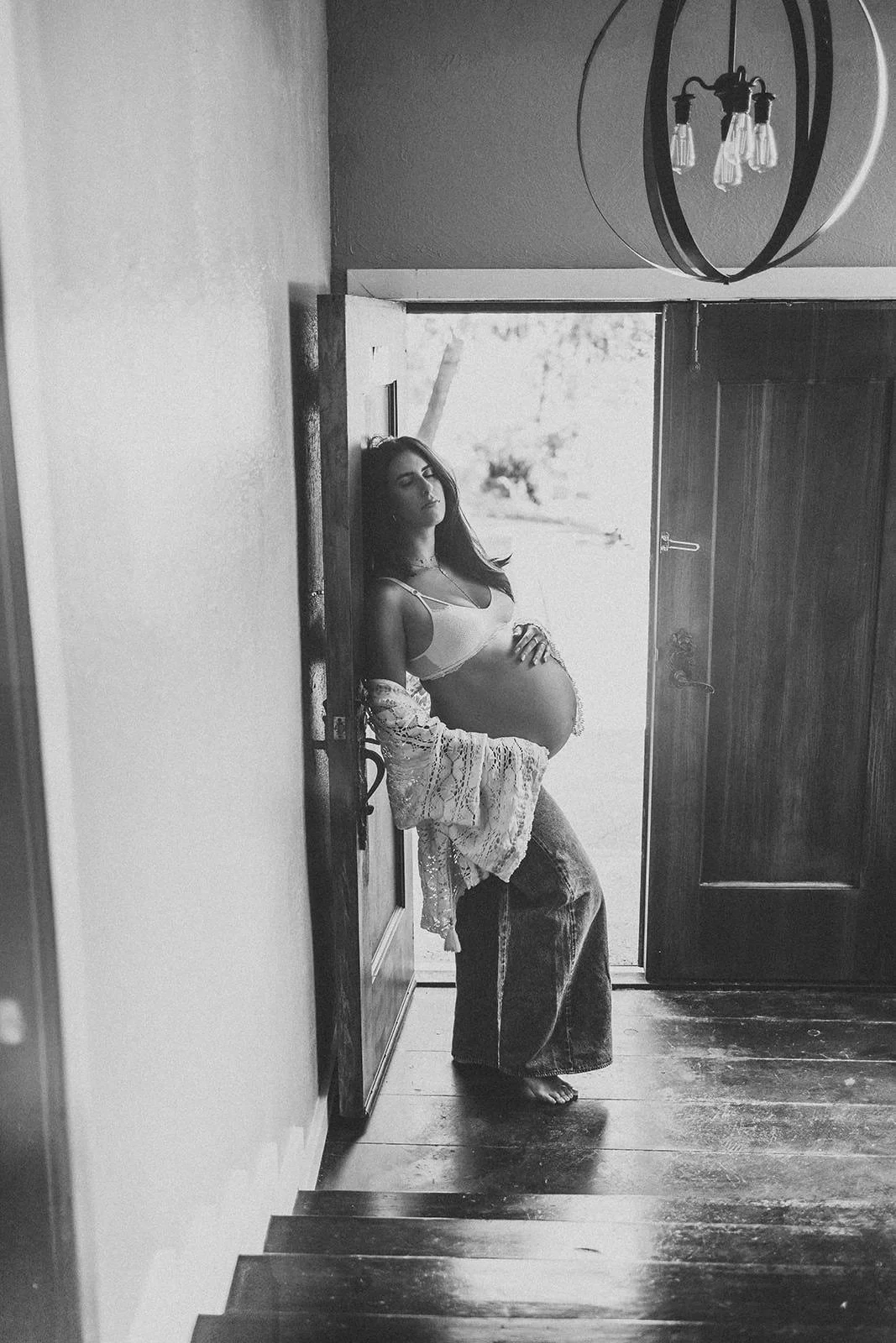 Black and white photo of a pregnant woman standing in a doorway at the top of stairs, wearing a bra and wide-leg jeans, with a lace robe draped over her shoulders, looking contemplative.