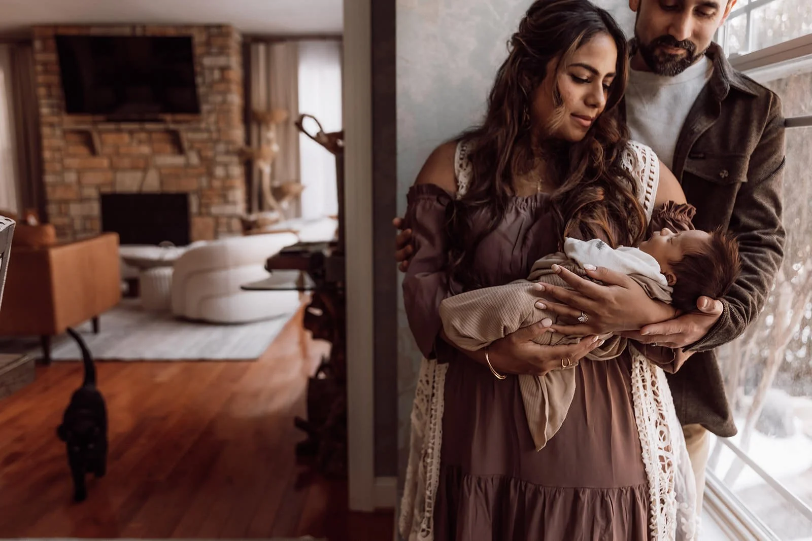 A woman and a man holding a newborn baby together near a window in a cozy home interior.