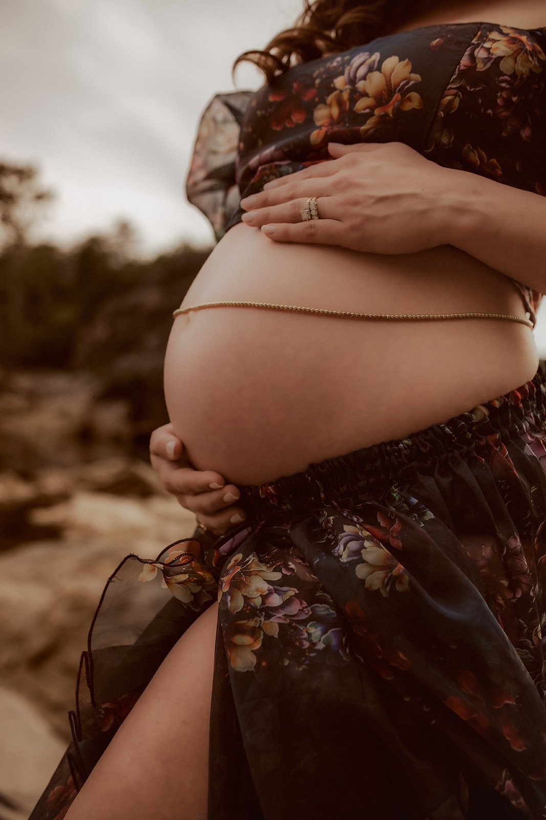 A pregnant woman outdoors, holding her belly with one hand, wearing a floral-patterned dress, with a gold belly chain visible.