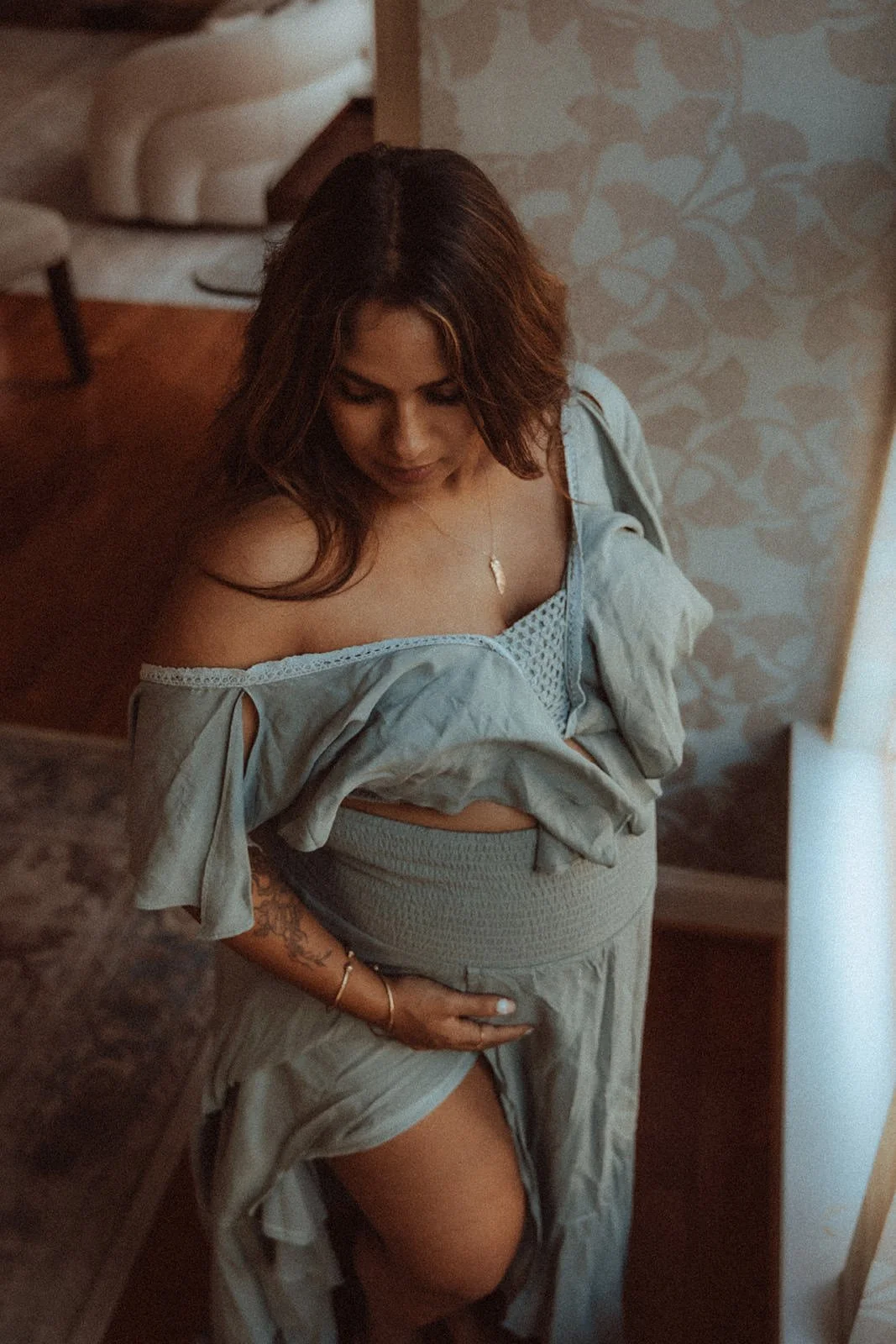 A woman with reddish-brown hair wearing a light blue off-shoulder top and a matching skirt, standing in a room with patterned wallpaper and a chair in the background, looking downward.