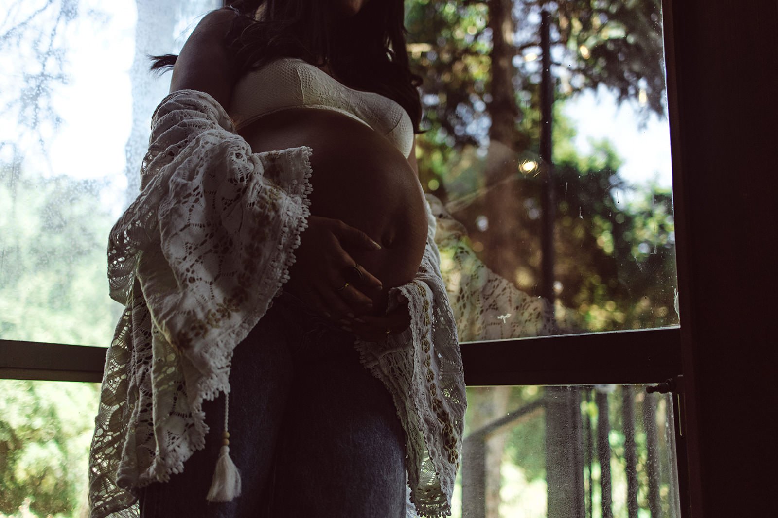 A pregnant woman standing indoors near a window, gently cradling her belly with one hand and standing in front of trees outside, seen through the window.