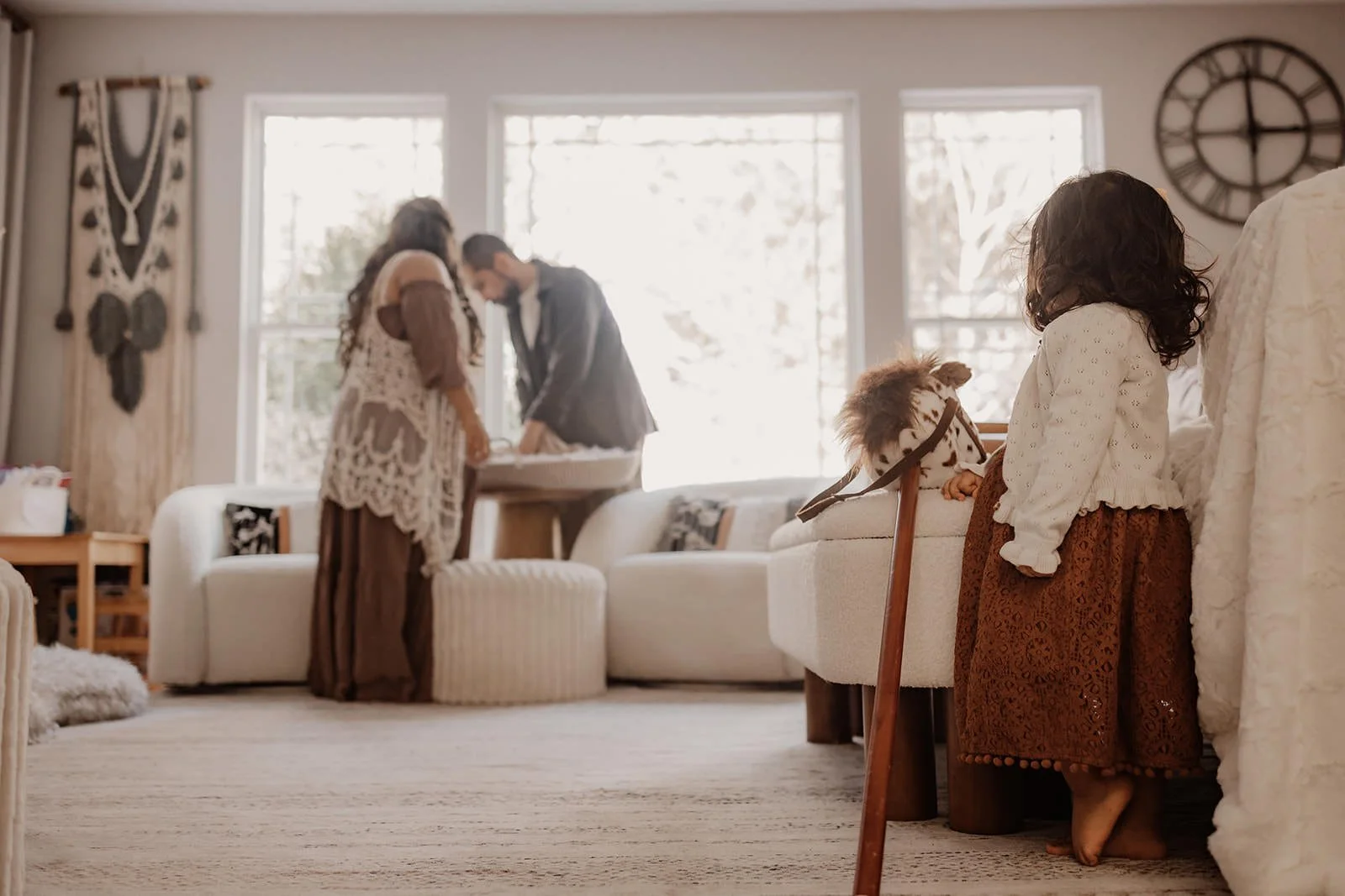 A young girl with dark curly hair wears a brown skirt and white sweater, standing by a white furniture piece with a small toy horse on it in a cozy living room with large windows and decorative wall art, while adults are in the background preparing or organizing something.