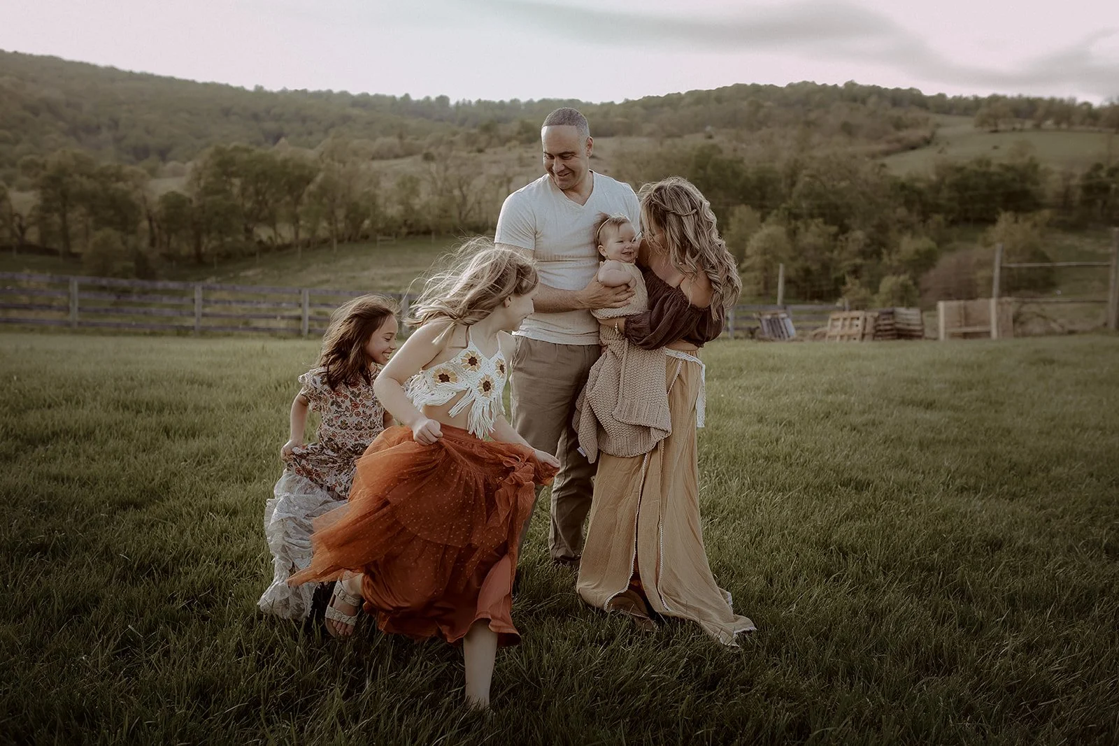 A family of five playing outdoors on green grass with hills and trees in the background, during daytime. They appear to be enjoying a joyful moment together.