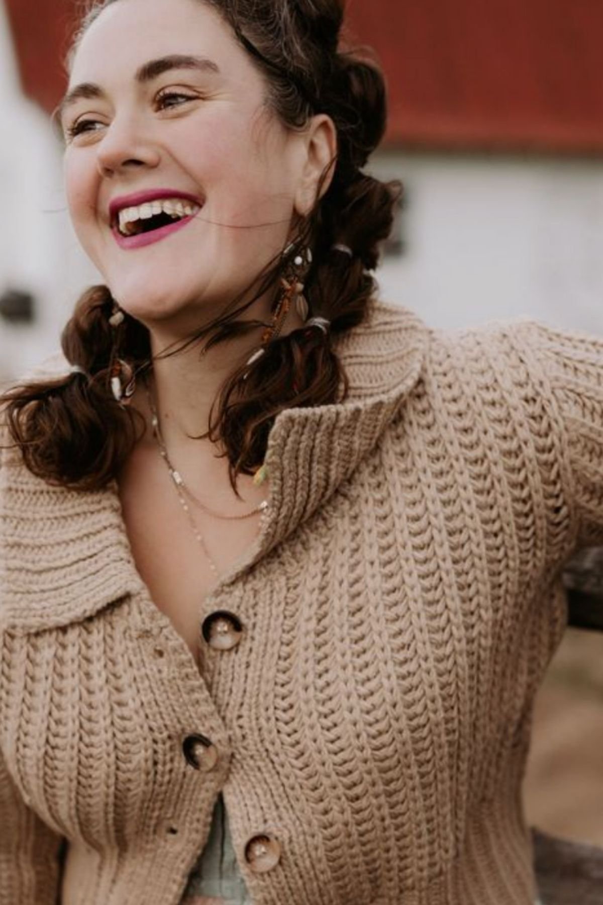Close-up of a smiling woman with curly brown hair and light skin, wearing a beige knit cardigan with large buttons and layered necklaces, outdoors with a blurred background.