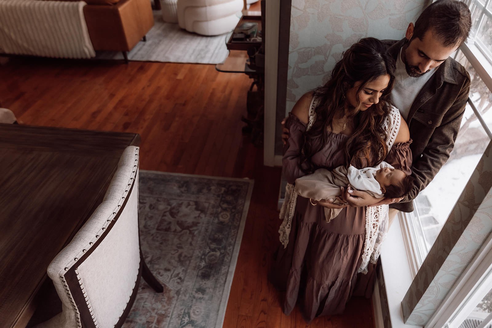 A couple holding a newborn baby by a large window in a cozy living room with hardwood floors, a patterned area rug, and a wooden dining table.