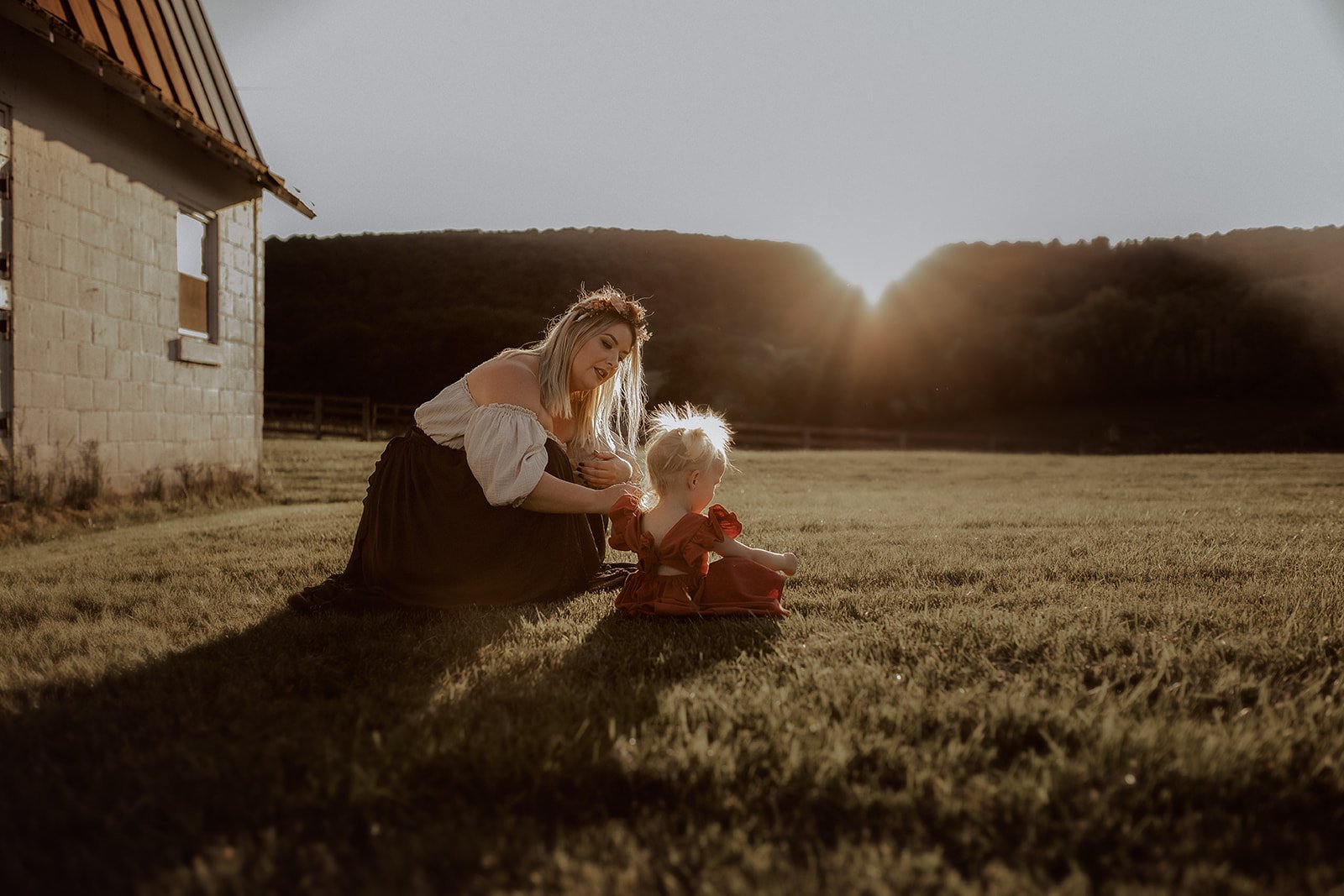 A woman and a young girl sit on the grass outside during sunset. The woman has blonde hair and wears a white off-the-shoulder top and a dark skirt. The girl has blonde hair in a bun and wears a red dress. They are enjoying a quiet moment together bes
