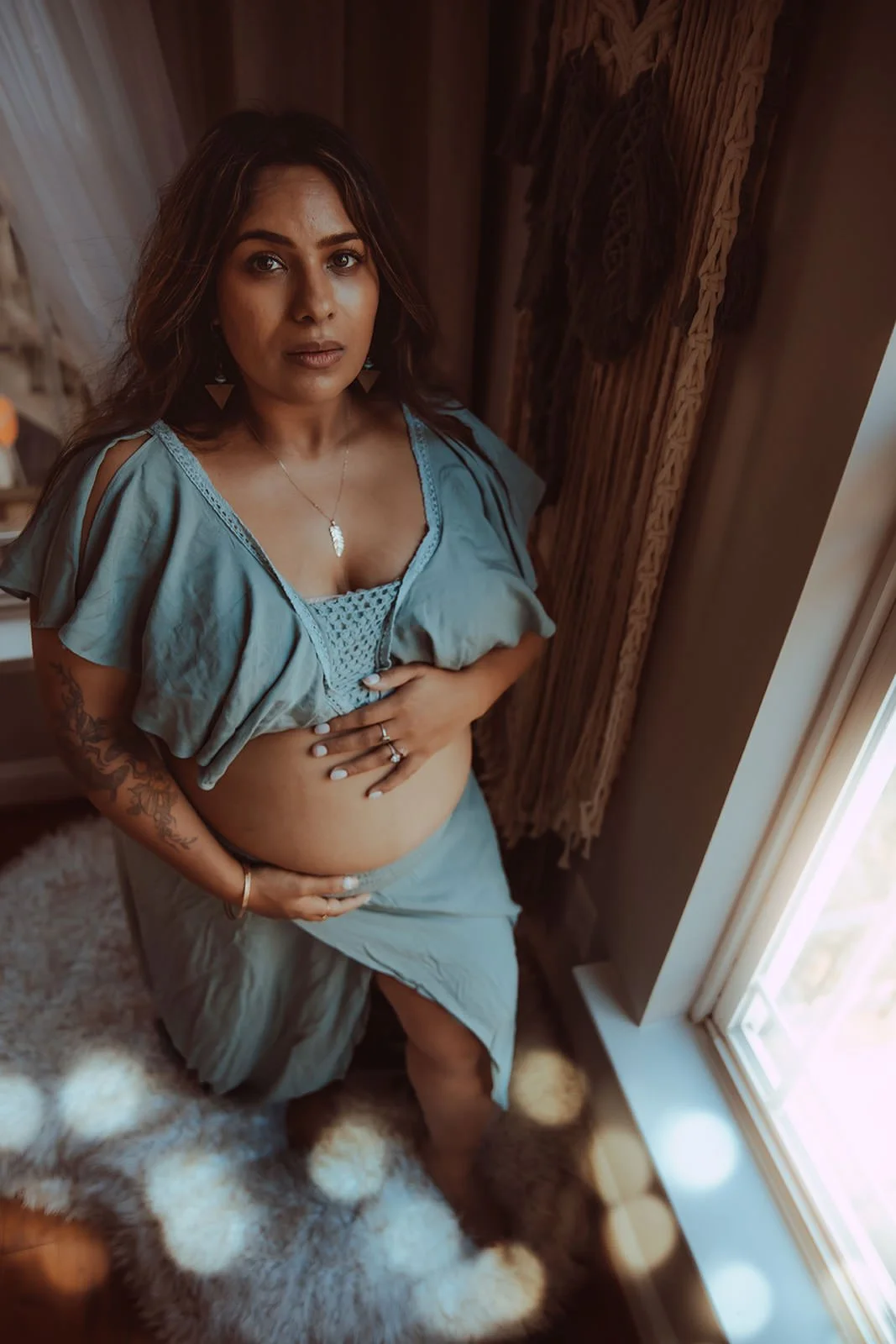 A woman with medium-length brown hair, wearing a light blue top and skirt, is standing by a window with natural light coming in, holding her pregnant belly, with a tattoo on her arm, jewelry, and looking at the camera.