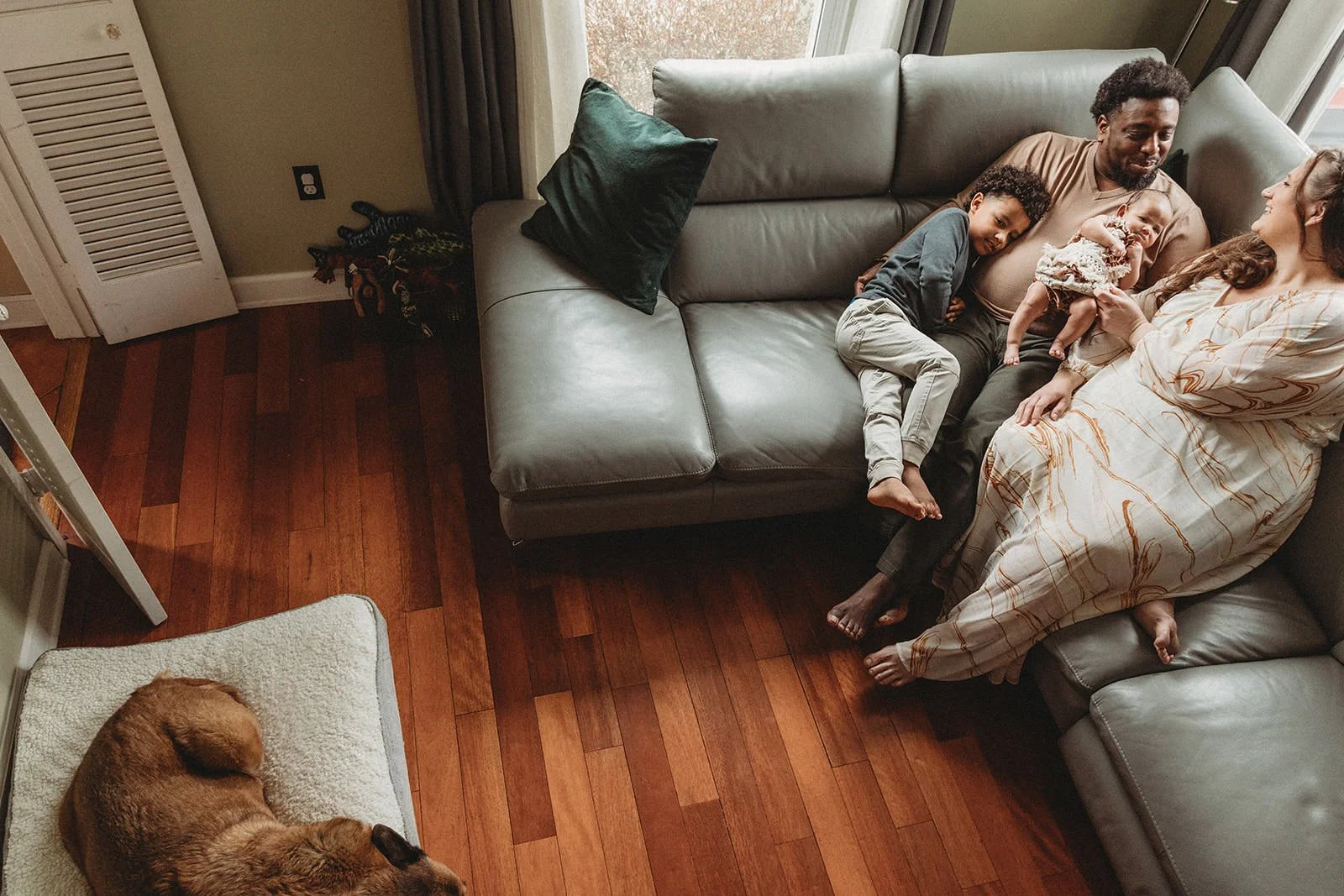 A family of four relaxing together on a gray sofa with a green pillow, next to a window with curtains. A woman in a patterned dress sits holding a baby girl, a man is sitting beside her with a boy leaning on his shoulder, all smiling and looking at e