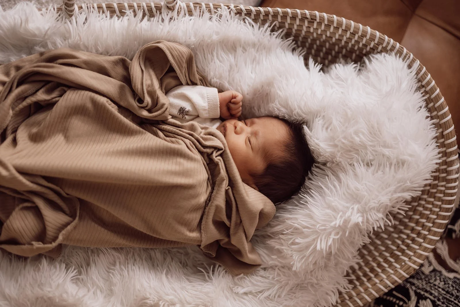 A sleeping baby in a beige and tan outfit, lying in a wicker bassinet with a white furry blanket lining.