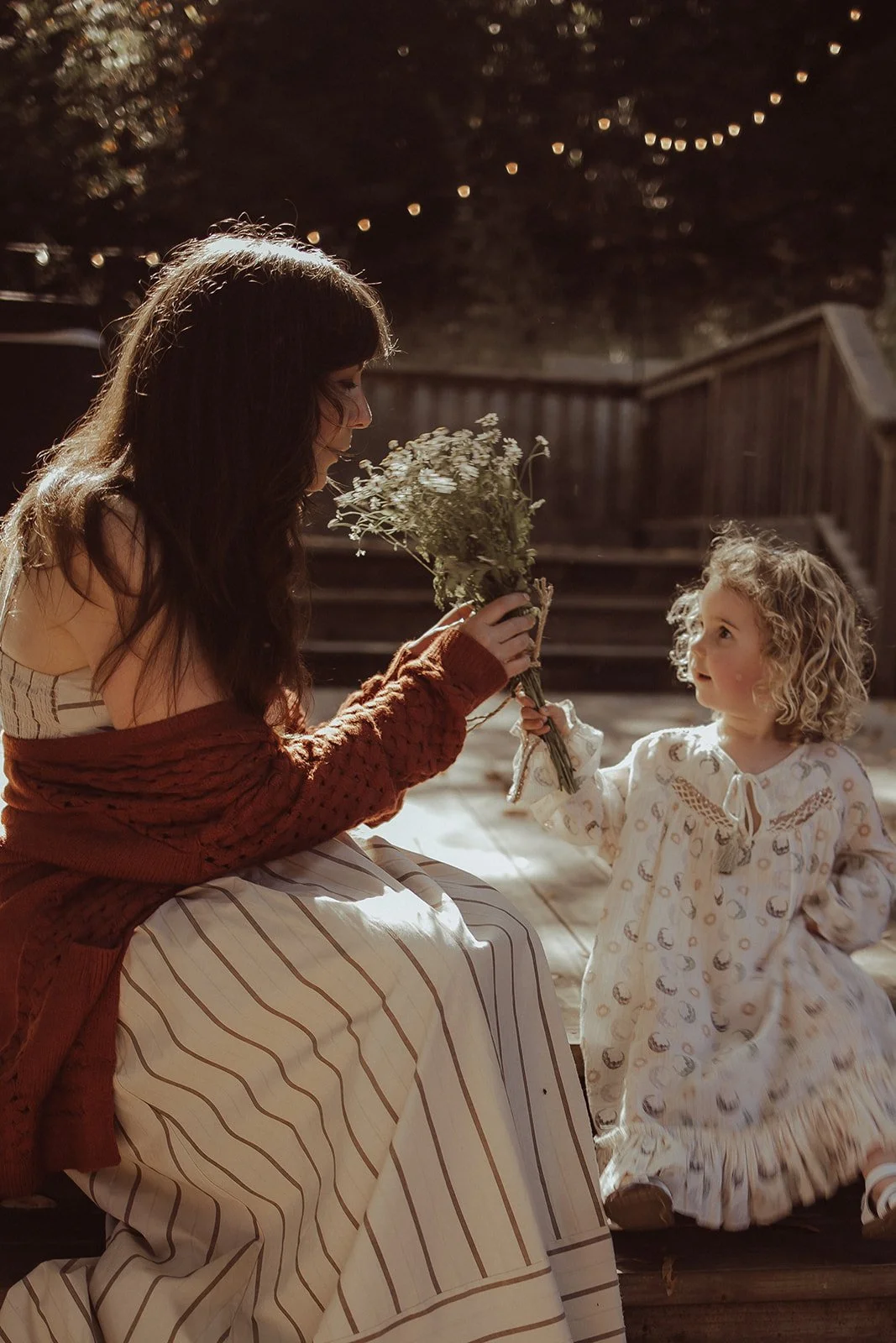 A woman sitting on stairs holding a bouquet of flowers, receiving one from a young girl with curly hair, dressed in a long patterned dress, in an outdoor setting at night.