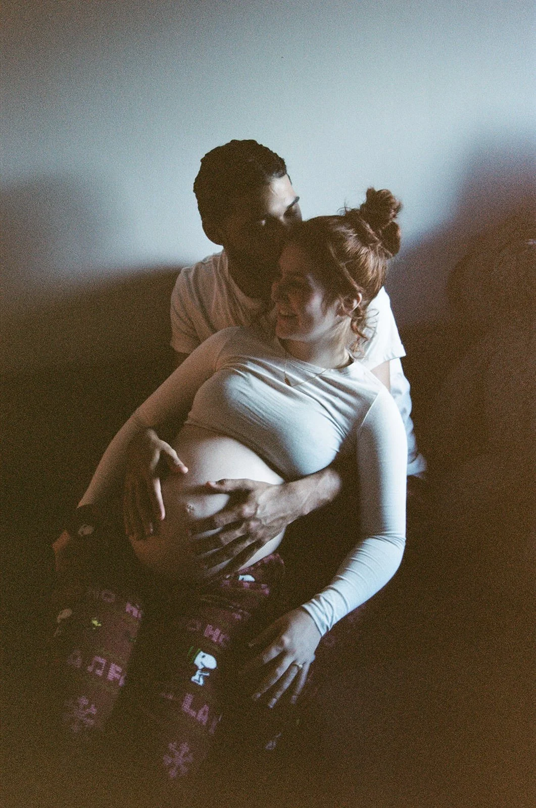 A couple sitting on a dark couch, embracing; the woman appears pregnant, and the man is holding her belly.